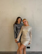 Frau mit blonden Haaren trägt kurzes Paillettenkleid in Gold und Frau mit brauen Haaren in silber 